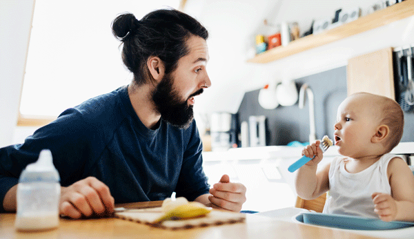 Mann schaut zu, wie Kleinkind am Esstisch versucht mit Gabel zu essen.