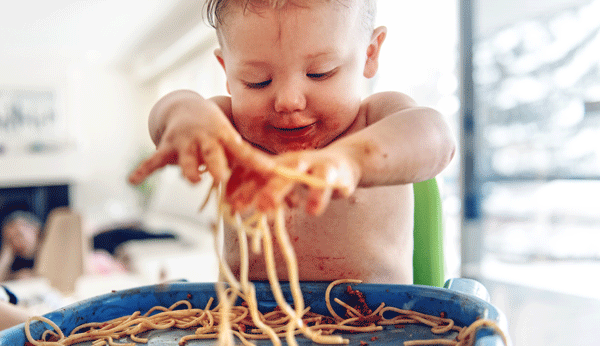 Kleinkind sitzt am Tisch, vollgeschmiert mit Tomatensauce, und spielt mit Spaghetti.