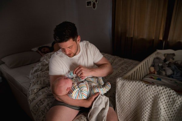 Vater sitzt auf Bettkante und hält kleines Baby mit Schnuller im Arm.