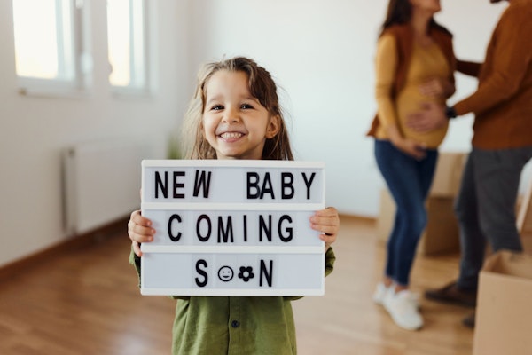 Ein Mädchen hält ein Schild hoch auf dem steht «New baby coming soon». Im Hintergrund hält der Mann den schwangeren Bauch der Frau.
