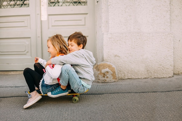 Kleines Mädchen und kleiner Junge sitzen auf Skateboard und rollen die Strasse hinunter.