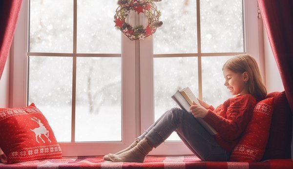 Mädchen liest ein Buch am weihnachtlich dekorierten Fenster.