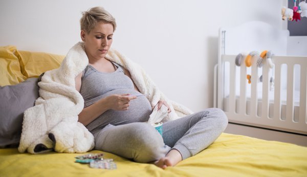 Krank in der Schwangerschaft: Schwangere Frau, eingekuschelt in Decke, kontrolliert auf Fieberthermometer ihre Temperatur. 