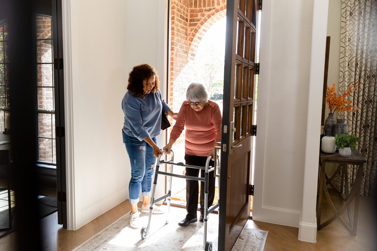 eine Pflegerin hilft einer Seniorin über die Türschwelle