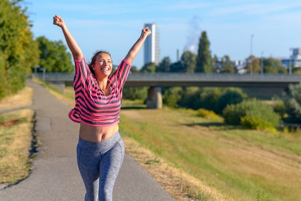 Frau mit Postpartum Body beim Joggen hebt freudig die Arme in die Luft