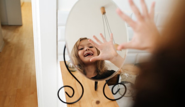 Ansicht von kleinem runden Spiegel, in dem das Gesicht von einem Mädchen mit Zahnlücke zu sehen ist, das sich selbst zuwinkt