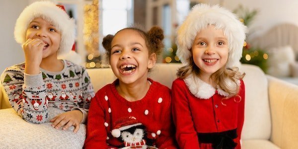 Drei lachende Kinder sitzen mit Popcorn auf dem Sofa. Sie tragen Weihnachtskleidung. 