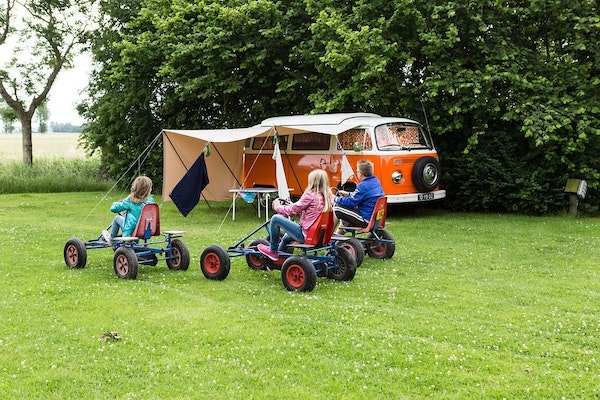 Drei Kinder auf Gokarts auf einer grünen Wiese, im Hintergrund steht ein alter VW-Bus mit Vorzelt