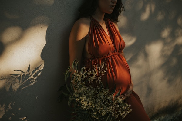 Eine schwangere Frau mit rotem Kleid lehnt sich an eine Wand an. Auf ihr ist der Schatten von Bäumen.