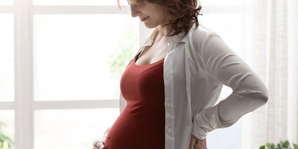 Schwangere Frau steht vor Fenster und hält sich eine Hand an den Bauch.