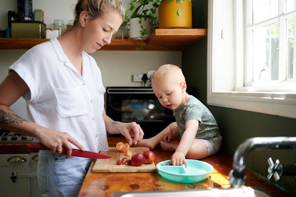 Junge Mutter beim Gemüse schneiden mit Kleinkind, das auf Küchenablage sitzt