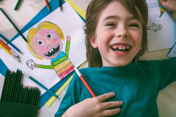 Junge liegt lachend auf dem Rücken am Boden neben einer Zeichnung von sich selbst mit einem Zahn in der Hand.