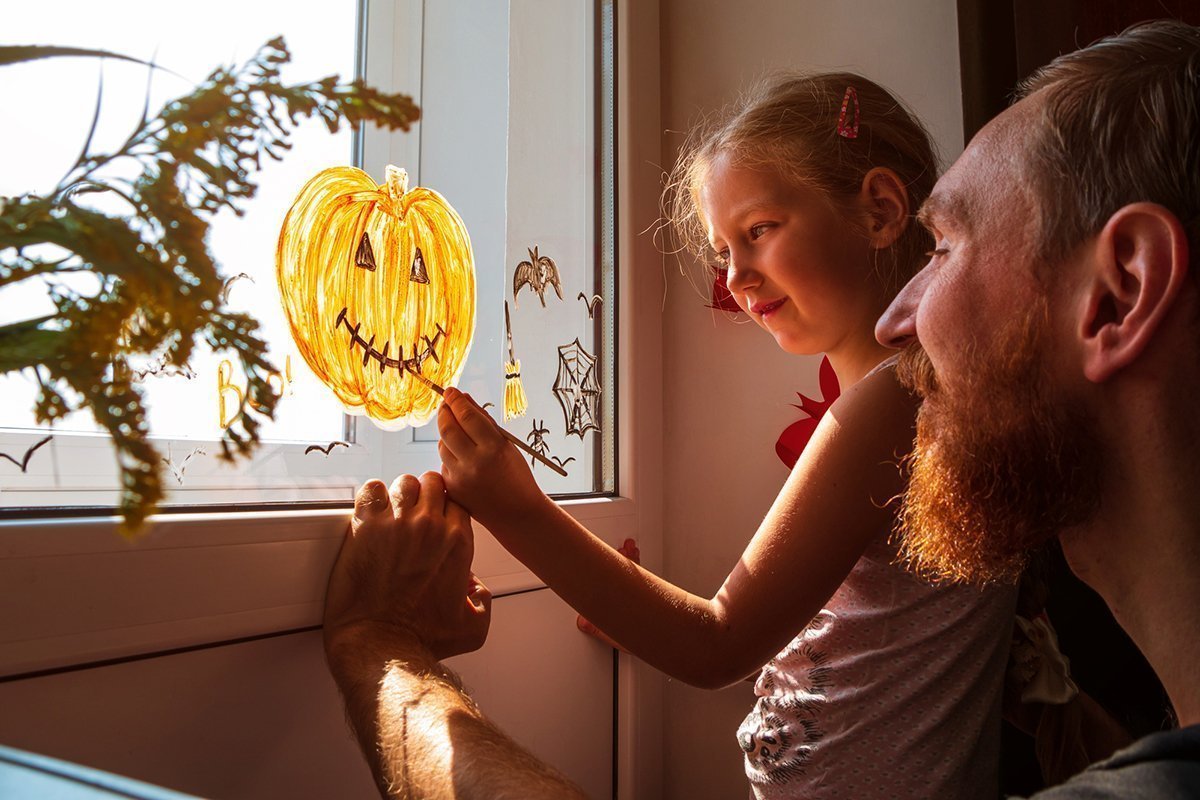 Kind mit Vater malt Kürbis auf Fenster zur Vorbereitung auf Halloween.