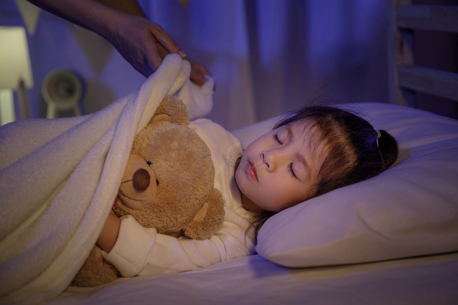 Mädchen mit Teddy im Arm wird im Bett zugedeckt