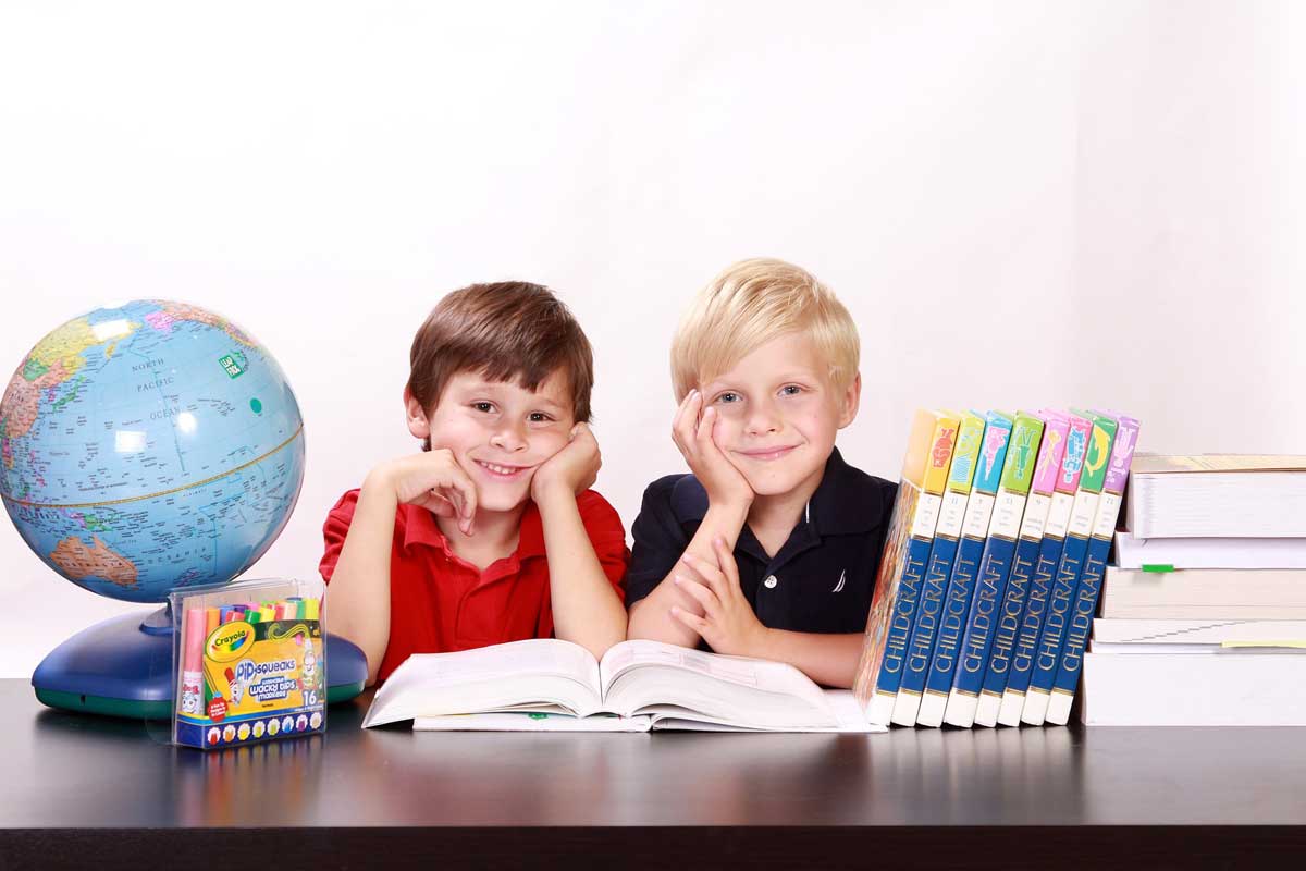 Zwei Kinder schauen in die Kamera. Vor sich auf dem Tisch haben sie Bücher, einen Globus und Stifte.