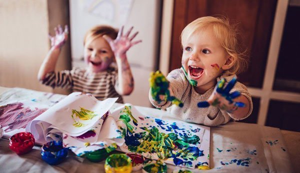Zwei kleine Mädchen beim Malen am Tisch, sie halten die farbverschmierten Hände in die Luft und lachen