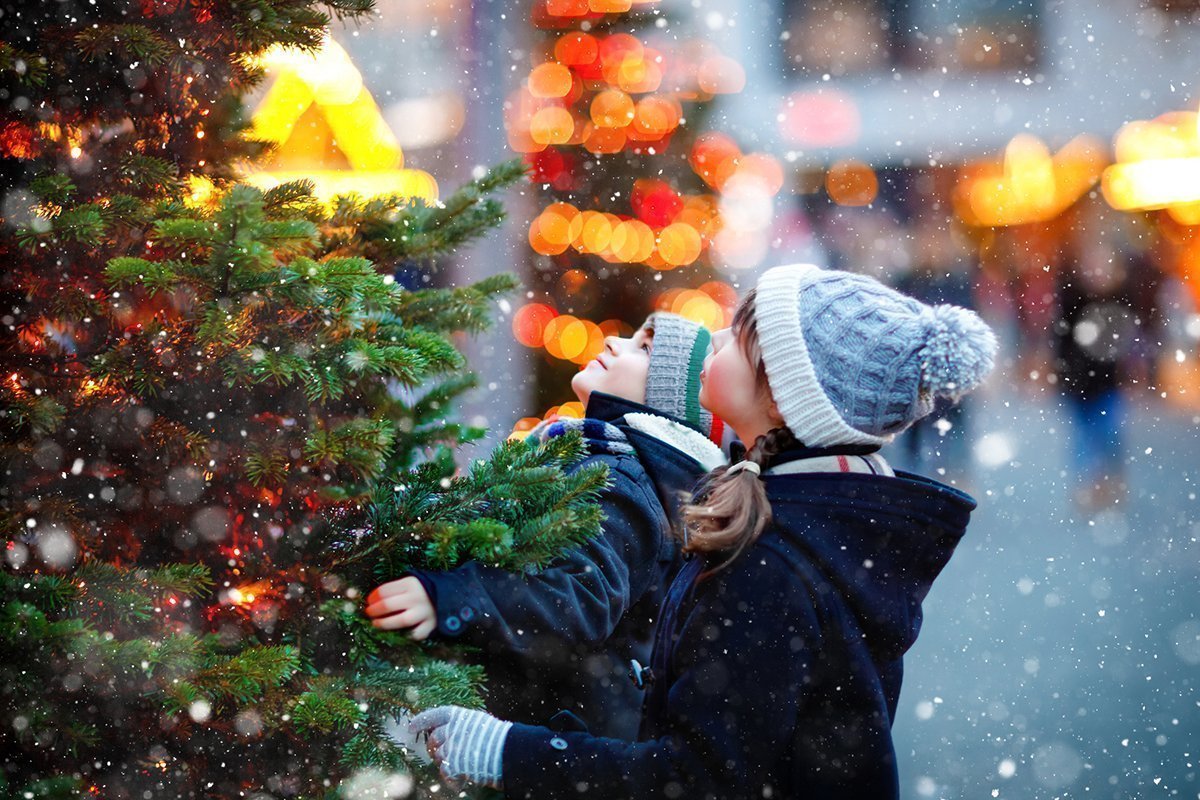 Zwei Kinder schauen sich glücklich einen Weihnachtsbau am Weihnachtsmarkt an, während es schneit.