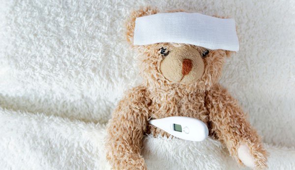 Teddybär mit Kompresse auf Stirn und Fieberthermometer unter Arm