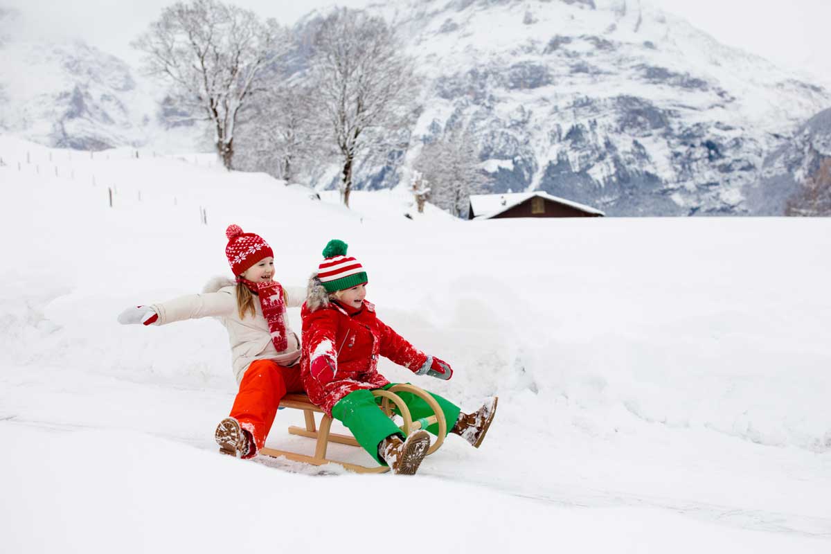 Zwei kleine Kinder schlitteln durch die verschneite Winterlandschaft.